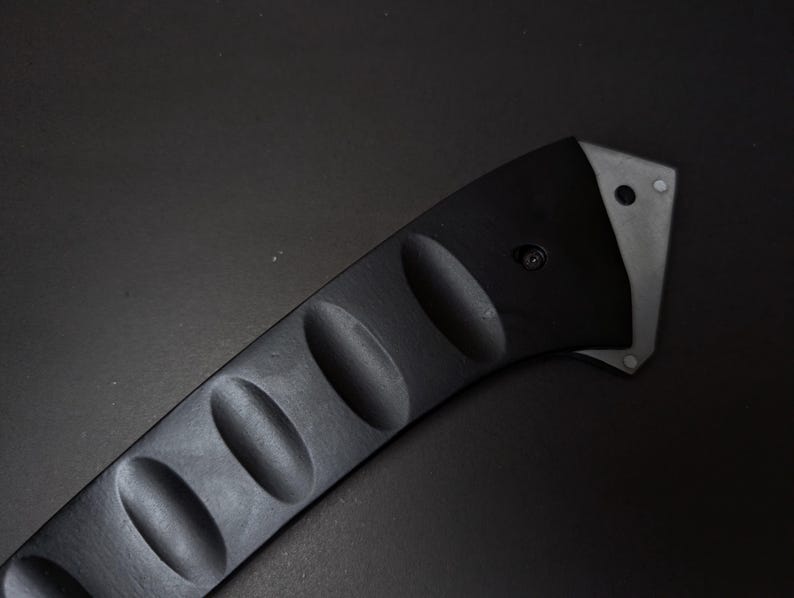May include: A black handle with a curved shape and four distinct oval indentations along its length. The handle features a metallic silver-toned end piece secured with a small black screw, showcasing a sleek and functional design for a tool.