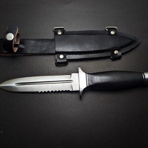May include: A stainless steel dagger with a serrated edge and black handle, next to a black leather sheath. The dagger has a double-edged blade and a silver crossguard. The sheath has a belt loop and is secured with metal rivets.