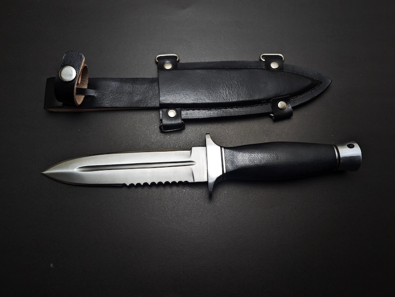 May include: A stainless steel dagger with a serrated edge and black handle, next to a black leather sheath. The dagger has a double-edged blade and a silver crossguard. The sheath has a belt loop and is secured with metal rivets.