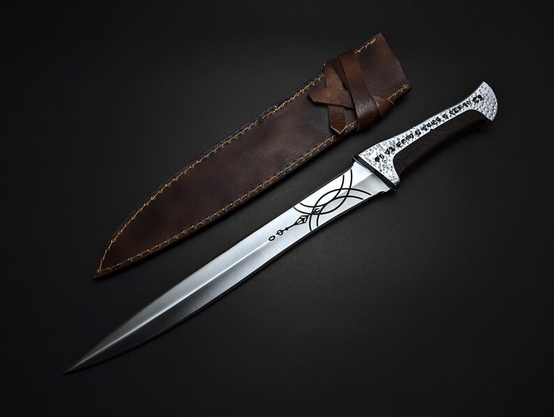 K&ouml;nnte beinhalten: Ein silberner Dolch mit einem verzierten Design und einem dunklen Holzgriff liegt neben einer braunen Lederscheide. Die Klinge hat eine scharfe Spitze und dekorative &Auml;tzungen. Die Scheide besteht aus braunem Leder mit einem gen&auml;hten Rand und einem Lederriemen.