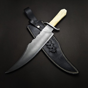 May include: A large, silver-bladed knife with a bone-colored handle rests in a black leather sheath. The blade has a serrated edge and a curved tip. The sheath is stitched with white thread and has a leather strap.