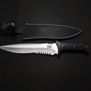 May include: A large, stainless steel knife with a black handle and a serrated edge. The knife is paired with a black leather sheath. The handle has gold-colored rivets. The knife is on a black surface.
