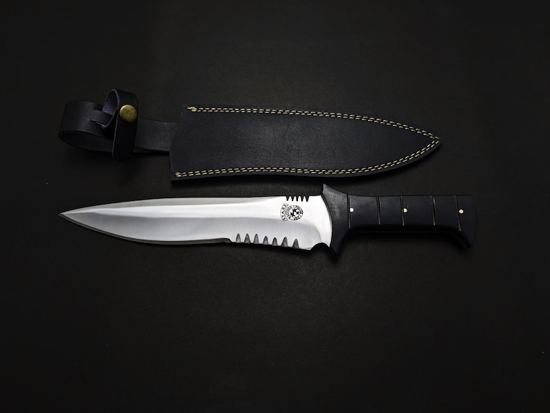 May include: A large, stainless steel knife with a black handle and a serrated edge. The knife is paired with a black leather sheath. The handle has gold-colored rivets. The knife is on a black surface.