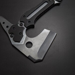 May include: A tactical hatchet featuring a matte gray metal head with a circular cutout and a sharp edge. The handle is constructed with black textured grip panels secured by visible screws, extending into a pointed spike at the top rear.