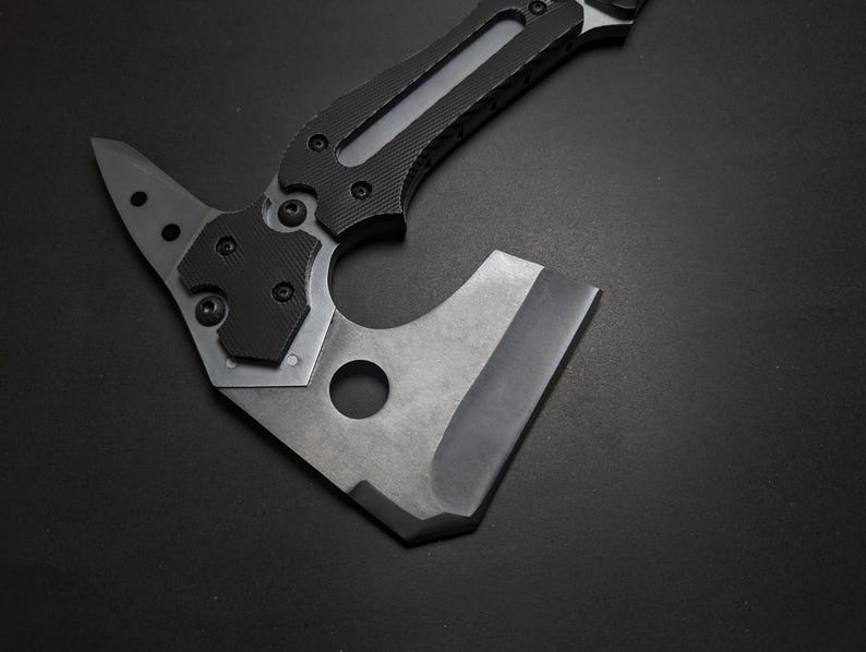 May include: A tactical hatchet featuring a matte gray metal head with a circular cutout and a sharp edge. The handle is constructed with black textured grip panels secured by visible screws, extending into a pointed spike at the top rear.