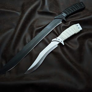 May include: Two knives on a dark brown surface. One is a long, black machete with a serrated edge and a textured handle. The other is a smaller knife with a silver blade and a white, wrapped handle.