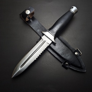 May include: A stainless steel dagger with a serrated edge and a black handle rests on top of a black leather sheath. The dagger is designed with a pointed tip and a silver metal bolster. The sheath has a strap with a snap closure.