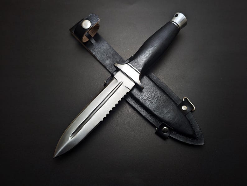 May include: A stainless steel dagger with a serrated edge and a black handle rests on top of a black leather sheath. The dagger is designed with a pointed tip and a silver metal bolster. The sheath has a strap with a snap closure.