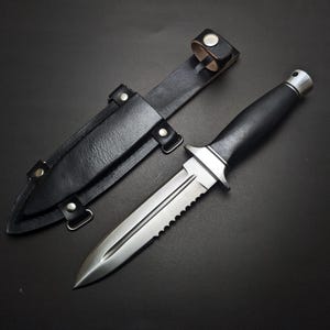 May include: A stainless steel knife with a black handle and a serrated edge, next to a black leather sheath. The knife has a pointed blade and a silver guard. The sheath has a belt loop and is secured with snaps.
