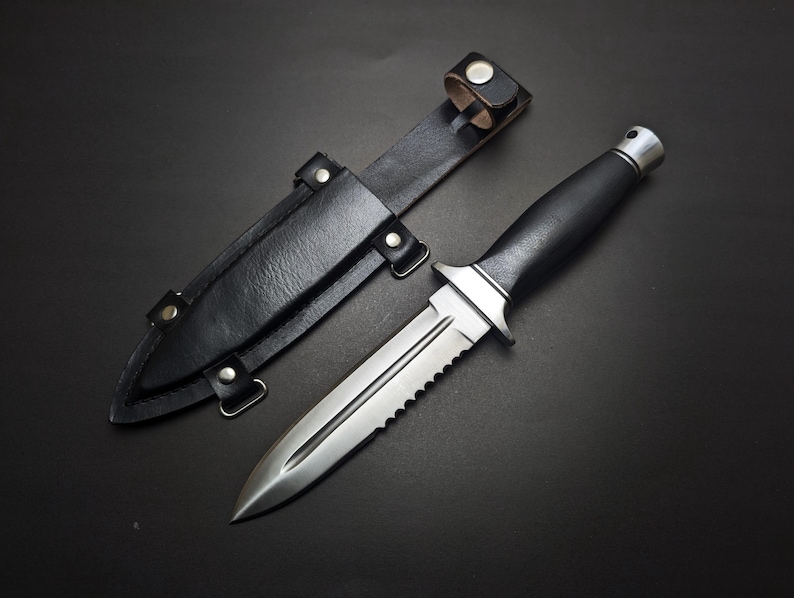May include: A stainless steel knife with a black handle and a serrated edge, next to a black leather sheath. The knife has a pointed blade and a silver guard. The sheath has a belt loop and is secured with snaps.