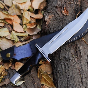 May include: A black-handled hunting knife with a silver blade and a brown leather sheath. The knife is lying on a tree trunk with fallen leaves around it.