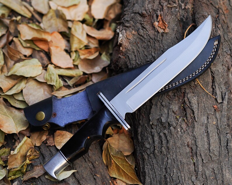 May include: A black-handled hunting knife with a silver blade and a brown leather sheath. The knife is lying on a tree trunk with fallen leaves around it.