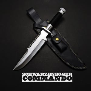 May include: A large, silver and black knife with a serrated edge, on a black leather sheath. The knife has a black handle and a silver blade. The text "SCHWARZENEGGER COMMANDO" is displayed below the knife.
