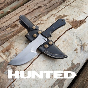 Puede incluir: Una funda de cuero negro con un cuchillo de hoja plateada con borde dentado. El cuchillo tiene un mango negro con detalles en latón. El cuchillo y la funda están sobre una superficie de madera. El texto "THE HUNTED" es visible en la imagen.