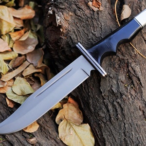 May include: A black-handled hunting knife with a silver blade and a black wooden handle. The knife is lying on a piece of wood with brown leaves scattered around it.