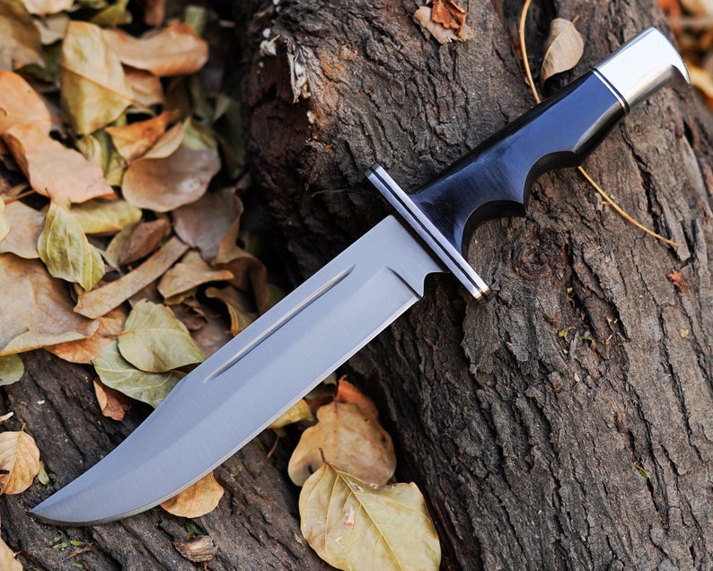 May include: A black-handled hunting knife with a silver blade and a black wooden handle. The knife is lying on a piece of wood with brown leaves scattered around it.
