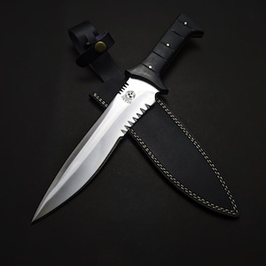 May include: A large, silver-bladed knife with a serrated edge and a black handle. The knife is paired with a black leather sheath. The handle has a series of rivets. The knife is approximately 12 inches long.
