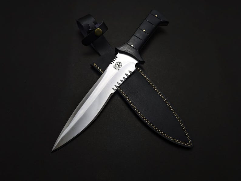 May include: A large, silver-bladed knife with a serrated edge and a black handle. The knife is paired with a black leather sheath. The handle has a series of rivets. The knife is approximately 12 inches long.