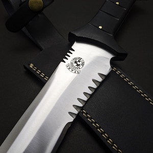May include: A close-up of a large, serrated-edge knife with a black handle and a leather sheath. The blade is silver with a logo that reads "S.T.A.R.S. Raccoon Police Department". The sheath is black with gold stitching.