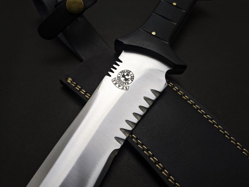 May include: A close-up of a large, serrated-edge knife with a black handle and a leather sheath. The blade is silver with a logo that reads "S.T.A.R.S. Raccoon Police Department". The sheath is black with gold stitching.