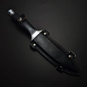 May include: A black dagger with a leather sheath. The dagger has a silver-colored metal handle and a black leather grip. The sheath is also black leather and has metal rivets and a belt loop.