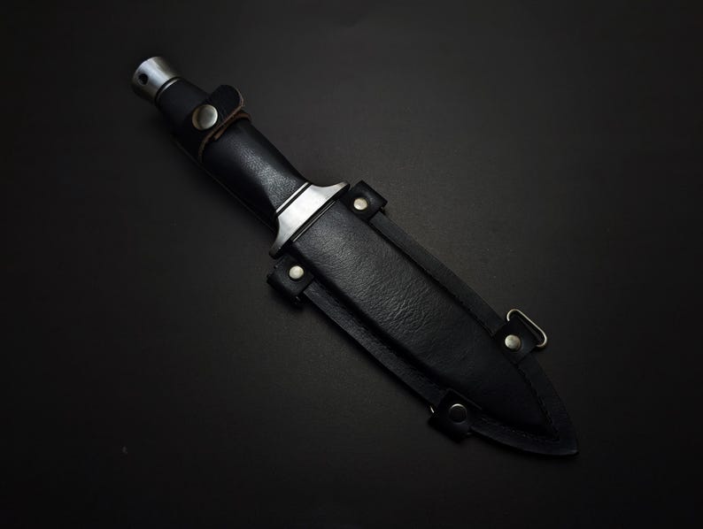 May include: A black dagger with a leather sheath. The dagger has a silver-colored metal handle and a black leather grip. The sheath is also black leather and has metal rivets and a belt loop.