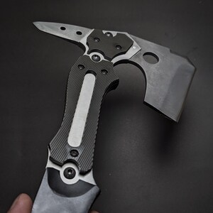 May include: A tactical multi-tool hatchet featuring a matte black finish, a sharp axe blade with a circular cutout, and a pointed spike on the opposite side. The handle is constructed with textured black grip panels and a central silver metal inlay.