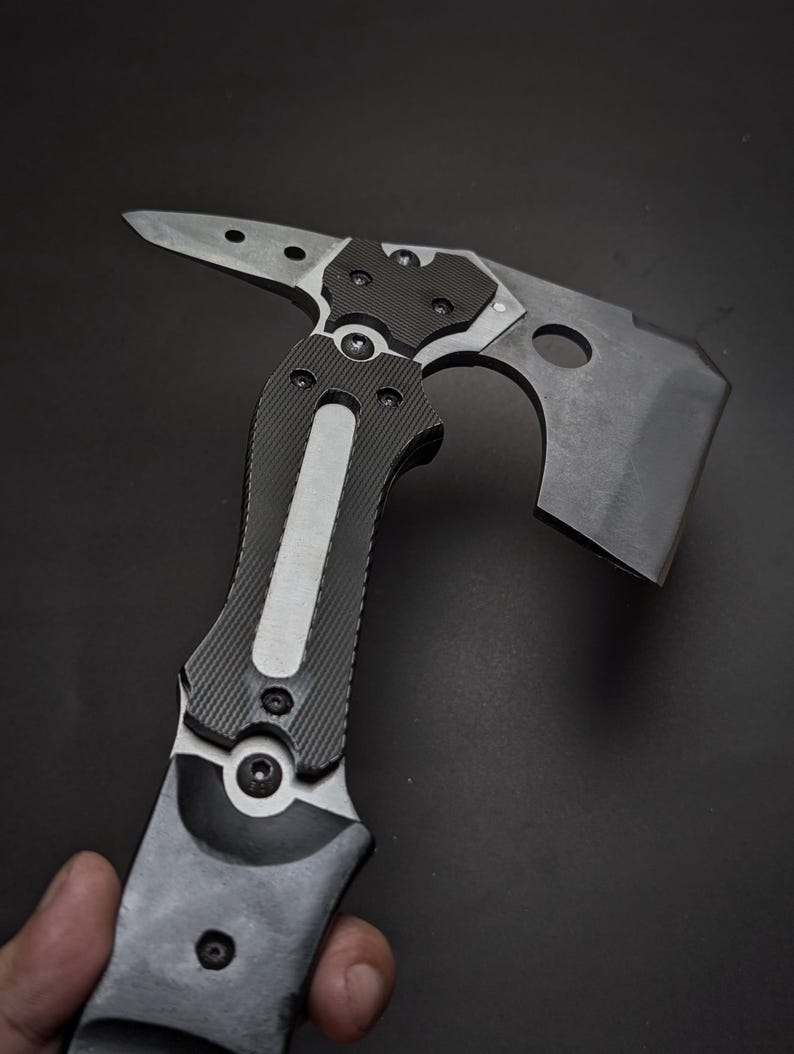 May include: A tactical multi-tool hatchet featuring a matte black finish, a sharp axe blade with a circular cutout, and a pointed spike on the opposite side. The handle is constructed with textured black grip panels and a central silver metal inlay.