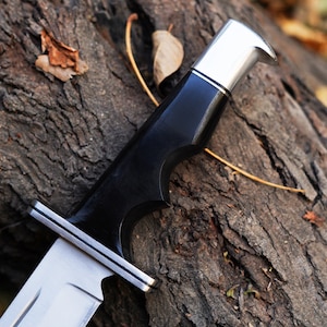 May include: Close-up of a black and silver hunting knife with a wooden handle. The knife is resting on a piece of tree bark.