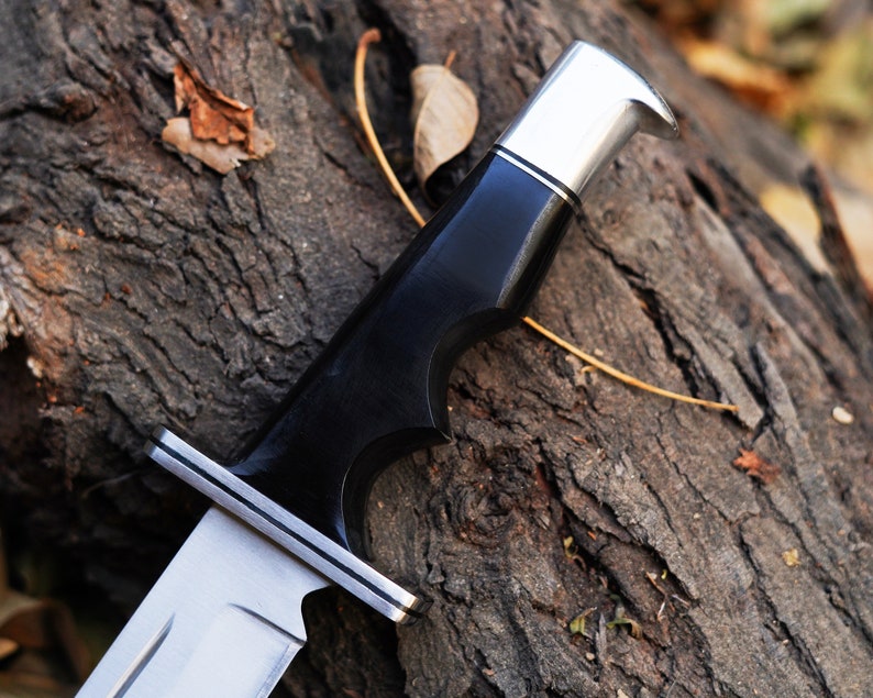 May include: Close-up of a black and silver hunting knife with a wooden handle. The knife is resting on a piece of tree bark.
