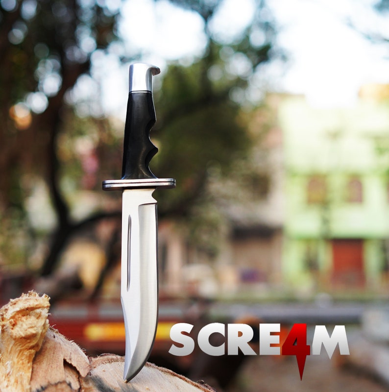 May include: A silver hunting knife with a black handle, standing upright on a wooden surface. The background is blurred and shows a green building with a red door. The text "SCREAM 4" is visible in the foreground.