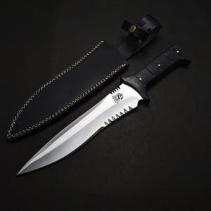 May include: A stainless steel dagger with a black handle and a serrated edge, accompanied by a black leather sheath. The dagger features a decorative emblem on the blade and is set against a dark background.