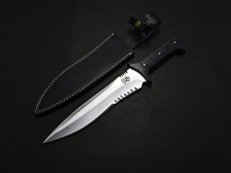 May include: A stainless steel dagger with a black handle and a serrated edge, accompanied by a black leather sheath. The dagger features a decorative emblem on the blade and is set against a dark background.