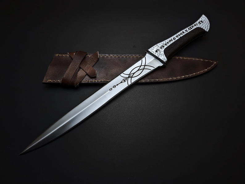 K&ouml;nnte beinhalten: Ein silberner Dolch mit kunstvollem Design auf Klinge und Griff, in einer braunen Lederscheide. Der Griff hat einen dunklen Holzgriff und einen dekorativen silbernen Knauf. Die Scheide hat einen Lederriemen.