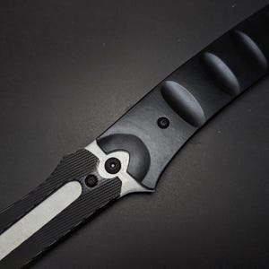 May include: A black and silver folding knife handle featuring a matte black finish with a series of rounded indentations along the spine. The handle includes a textured black grip section and a silver metal bolster secured by two small black screws.