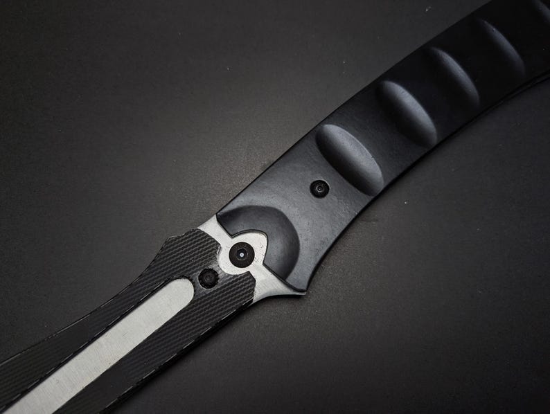May include: A black and silver folding knife handle featuring a matte black finish with a series of rounded indentations along the spine. The handle includes a textured black grip section and a silver metal bolster secured by two small black screws.