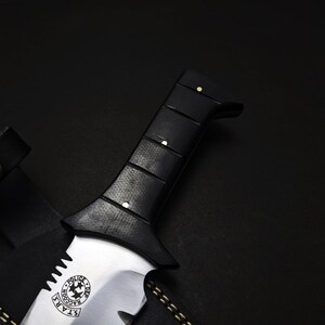 May include: Close-up of a black-handled knife with a silver blade. The handle has a textured grip and is secured with small rivets. The blade features a circular emblem with text and stars. A black leather sheath is partially visible.