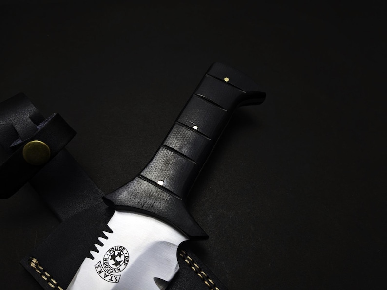 May include: Close-up of a black-handled knife with a silver blade. The handle has a textured grip and is secured with small rivets. The blade features a circular emblem with text and stars. A black leather sheath is partially visible.