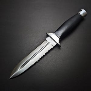May include: A stainless steel knife with a black handle and a serrated edge. The knife has a double-edged blade and a silver metal guard. The handle is made of a textured black material. The knife is set against a black background.