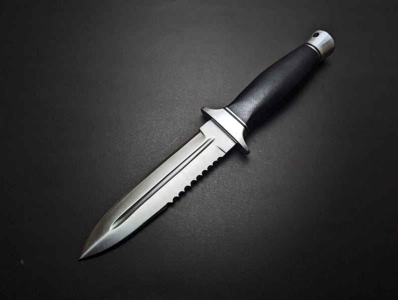 May include: A stainless steel knife with a black handle and a serrated edge. The knife has a double-edged blade and a silver metal guard. The handle is made of a textured black material. The knife is set against a black background.