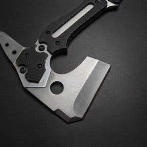 May include: A tactical hatchet featuring a silver-toned metal blade with a circular cutout and a pointed rear spike. The handle is constructed with black textured grip panels secured by multiple screws, extending into a metallic frame.