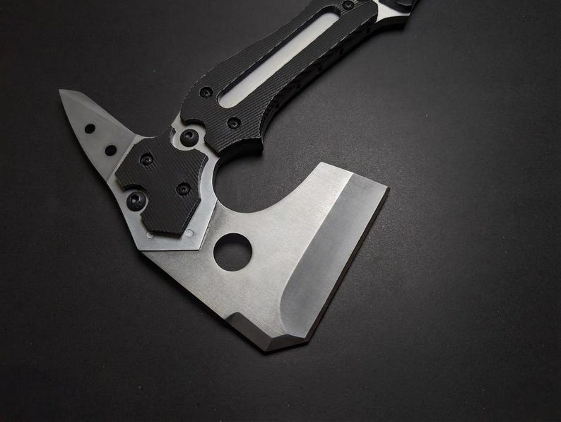 May include: A tactical hatchet featuring a silver-toned metal blade with a circular cutout and a pointed rear spike. The handle is constructed with black textured grip panels secured by multiple screws, extending into a metallic frame.