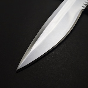 May include: Close-up of a stainless steel knife blade. The blade is sharp and pointed, with a polished finish. The knife appears to be a hunting or survival knife, with a serrated edge near the handle. The background is black.