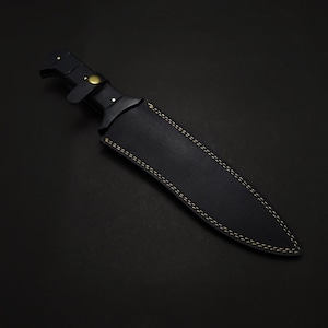 May include: A black knife with a leather sheath. The knife handle has a brass-colored rivet and a leather strap. The sheath is black with yellow stitching. The knife is on a black background.