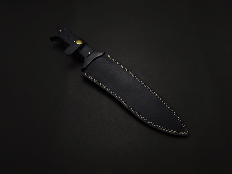 May include: A black knife with a leather sheath. The knife handle has a brass-colored rivet and a leather strap. The sheath is black with yellow stitching. The knife is on a black background.