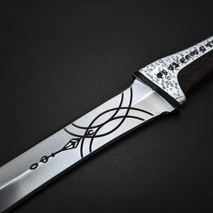 K&ouml;nnte beinhalten: Nahaufnahme eines silber-schwarzen Messers mit aufwendigen &Auml;tzmustern. Die Klinge weist eine Reihe geschwungener Linien und ein dekoratives Muster auf. Der Griff ist dunkel mit strukturierter Oberfl&auml;che, der Knauf ist mit kleinen, kreisf&ouml;rmigen Details verziert.