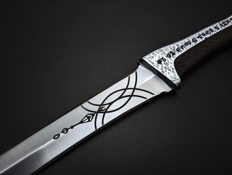K&ouml;nnte beinhalten: Nahaufnahme eines silber-schwarzen Messers mit aufwendigen &Auml;tzmustern. Die Klinge weist eine Reihe geschwungener Linien und ein dekoratives Muster auf. Der Griff ist dunkel mit strukturierter Oberfl&auml;che, der Knauf ist mit kleinen, kreisf&ouml;rmigen Details verziert.