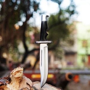 May include: A silver and black hunting knife with a black handle and a silver blade. The knife is standing upright on a piece of wood.