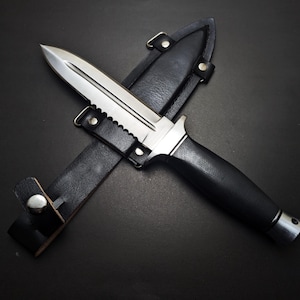 May include: A stainless steel dagger with a serrated edge and black handle rests in its black leather sheath. The knife has a double-edged blade and a silver metal bolster. The sheath has a belt loop and a snap closure.