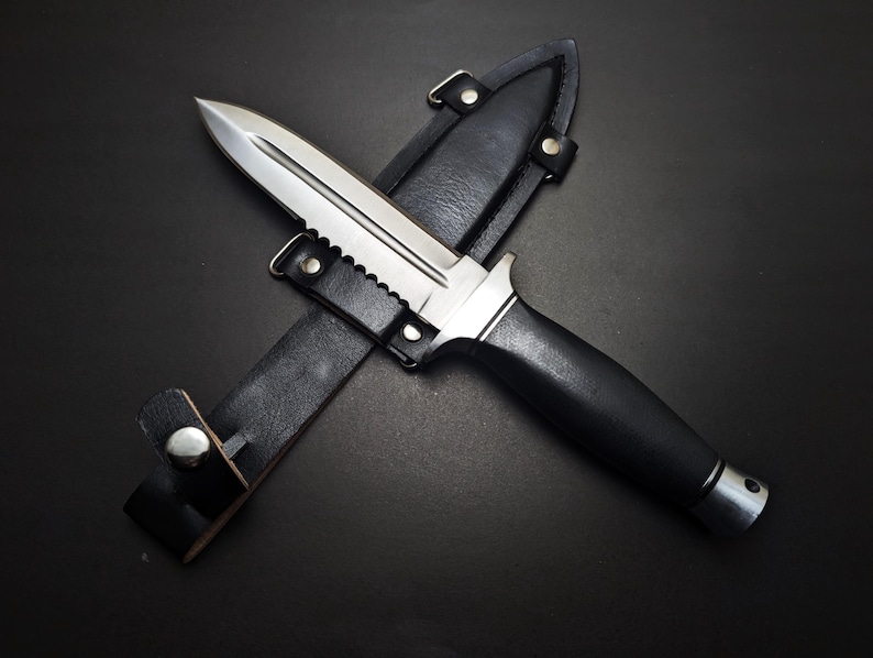 May include: A stainless steel dagger with a serrated edge and black handle rests in its black leather sheath. The knife has a double-edged blade and a silver metal bolster. The sheath has a belt loop and a snap closure.
